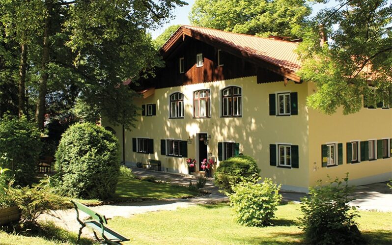 Farbfotografie eines traditionellen bayerischen Hauses mit gelber Fassade, dunkelgrünen Fensterläden und einem Dach mit roten Ziegeln. Das Gebäude ist von großen Bäumen und gepflegten Gärten umgeben, die einen schattigen und ruhigen Eindruck vermitteln. Im Vordergrund stehen ein Weg und eine Holzbank, die zur entspannten Atmosphäre beitragen. Die Szene wirkt sonnig und idyllisch, typisch für ein ländliches Anwesen.