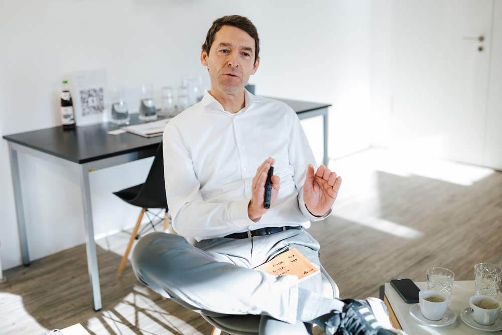 Ein Mann in einem hellen Raum sitzt entspannt mit übergeschlagenen Beinen und spricht gestikulierend. Auf dem Tisch im Hintergrund stehen Getränke und Notizen. Die Atmosphäre wirkt professionell und freundlich.