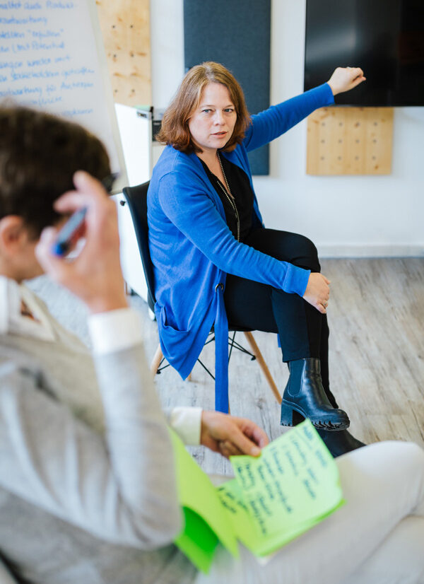 Eine Frau in einem blauen Cardigan gestikuliert energisch während eines Workshops und richtet ihren Blick auf jemanden außerhalb des Bildes. Im Vordergrund sitzt eine weitere Person, die Notizen in grünen Blättern hält. Im Hintergrund sind ein Flipchart und eine dunkle Wand sichtbar, was den Raum als Seminarumgebung kennzeichnet.