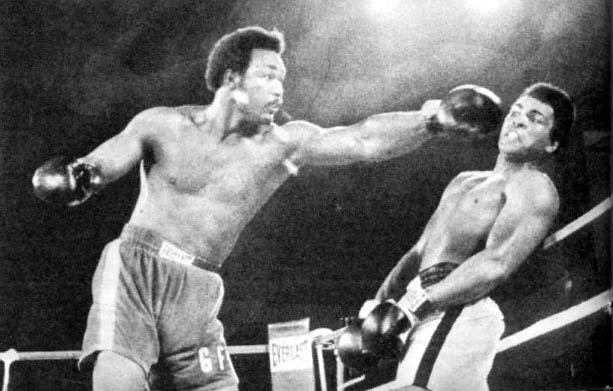 Schwarz-Weiß-Fotografie eines Boxkampfs zwischen George Foreman und Muhammad Ali. Foreman, links im Bild, wirft einen kräftigen Schlag, während Ali, rechts, dem Treffer geschickt ausweicht. Beide Boxer sind mit freiem Oberkörper im Ring, und die Szene zeigt die Intensität des berühmten Kampfes, bekannt als 'Rumble in the Jungle' (1974), bei dem Ali schließlich durch K.o. gewann.