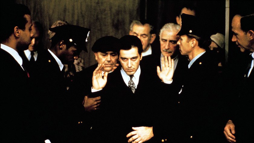 Filmszene aus 'Der Pate II' (1974), in der Michael Corleone, gespielt von Al Pacino, inmitten einer Gruppe von Männern und Polizisten zu sehen ist. Michael hebt seine Hände in einer defensiven Geste, während er einen ernsten und konzentrierten Gesichtsausdruck zeigt. Die Szene vermittelt eine angespannte Atmosphäre und symbolisiert die zunehmende Isolation und Machtkämpfe, die die Figur durchlebt. Die umstehenden Männer, teils in Uniform, verstärken den Eindruck von Bedrohung und Verhör.