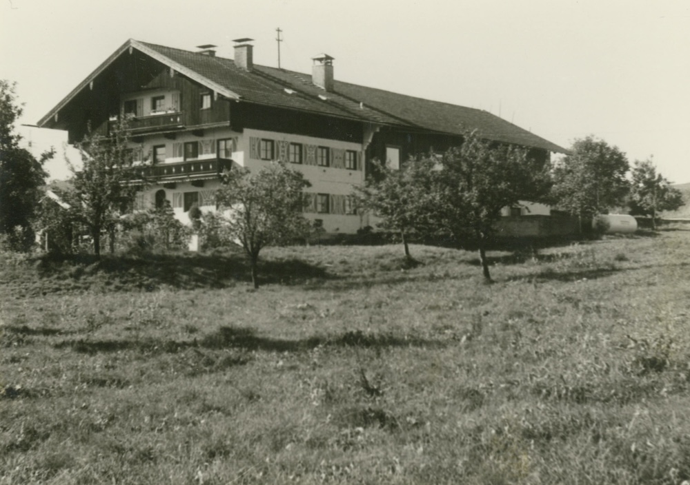 Schwarz-Weiß-Fotografie eines traditionellen bayerischen Bauernhauses mit Satteldach und Holzfassade. Das Gebäude hat zahlreiche Fensterläden und einen Balkon. Im Vordergrund befindet sich eine Wiese mit einzelnen Obstbäumen. Das Bild vermittelt eine ländliche, ruhige Atmosphäre und scheint aus den 1970er Jahren zu stammen.