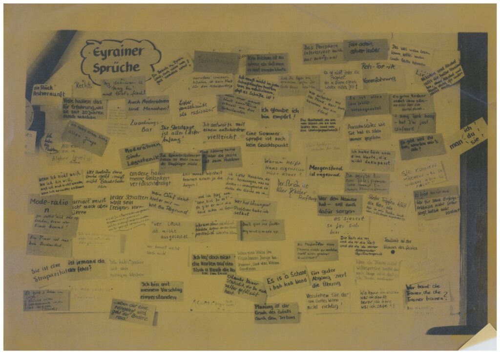 Ein großformatiges Poster mit der Überschrift 'Eyrainer Sprüche', auf dem viele handgeschriebene Zettel und Notizen angepinnt sind. Die Zettel enthalten verschiedene Zitate, Aussagen und humorvolle Sprüche, die in unterschiedlicher Schriftgröße und -art verfasst sind. Einige Notizen sind verblasst, andere heben sich deutlicher ab. Das Gesamtbild wirkt wie eine Sammlung von persönlichen Anekdoten und beliebten Redewendungen aus verschiedenen Kontexten.