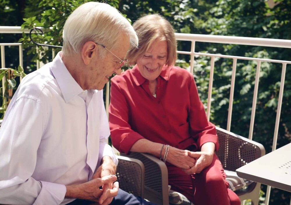 Das Gründerpaar der Firma ComTeam sitzt lächelnd auf einer Terrasse. Walther Straub trägt ein weißes Hemd und hat weißes Haar, Heide Straub trägt eine rote Bluse und schaut lächelnd auf ihn. Sie sitzen entspannt nebeneinander, umgeben von Pflanzen im Hintergrund, die eine sommerliche, freundliche Atmosphäre schaffen.